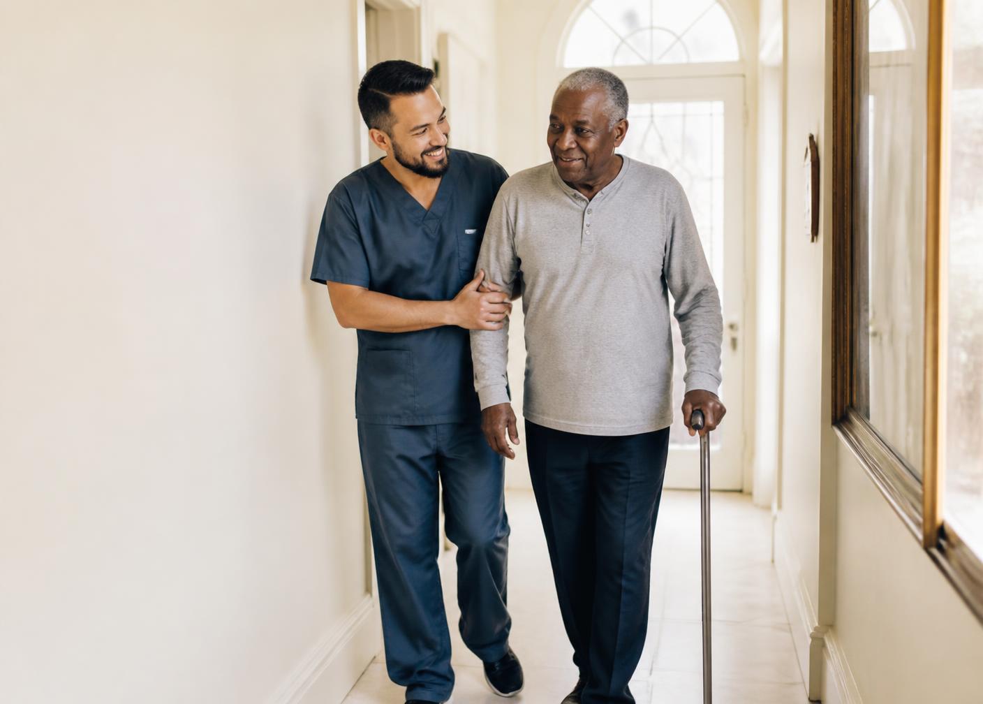 A caregiver supporting an older gentleman as he walks with a cane at home